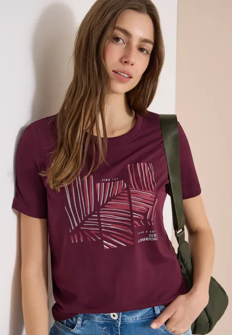 Frau trägt weinrotes T-Shirt mit abstraktem Blattmuster und grünem Rucksack