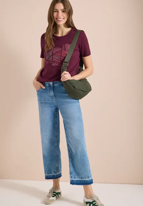 Frau in lila T-Shirt mit Muster, weiter Jeans, grüner Umhängetasche und Sneakern.