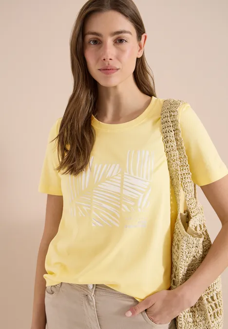 Vrouw in een geel t-shirt met wit grafisch dessin, beige broek en een gehaakte tas over de schouder.