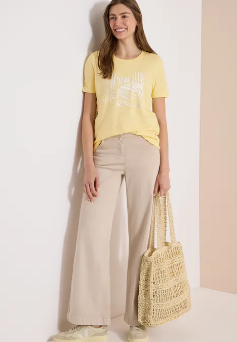 Persoon draagt een geel T-shirt, beige wijde broek en houdt een gehaakte boodschappentas vast.