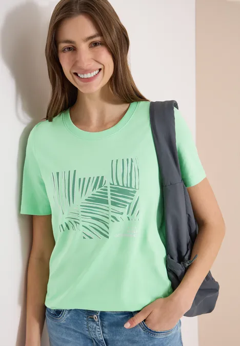 Frau in grünem T-Shirt mit Blattmuster, eine Hand in der Tasche, Rucksack über der Schulter.