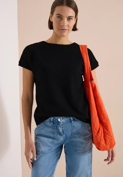 Eine Person trägt ein schwarzes T-Shirt, blaue Jeans und eine orangefarbene gesteppte Umhängetasche.