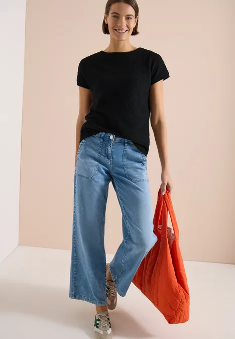 Frau in schwarzem T-Shirt, weitem Jeans und orangefarbener Tasche vor hellem Hintergrund.