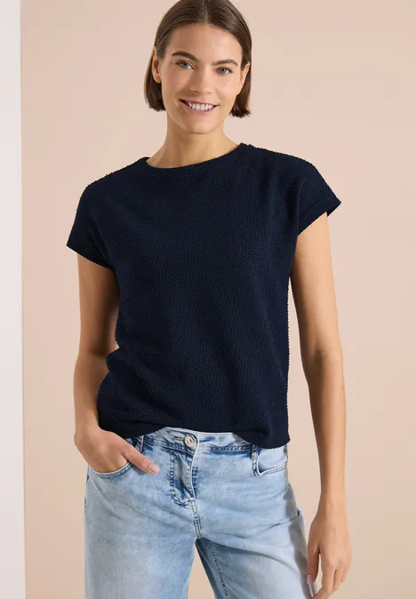 Frau trägt dunkelblaues T-Shirt mit kurzen Ärmeln und helle Jeans vor beigem Hintergrund.