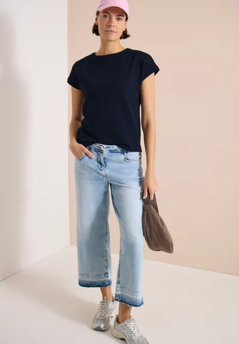 Frau in schwarzem T-Shirt, hellblauen Kickflare-Jeans, silbernen Sneakern und Taupe-Umhängetasche.