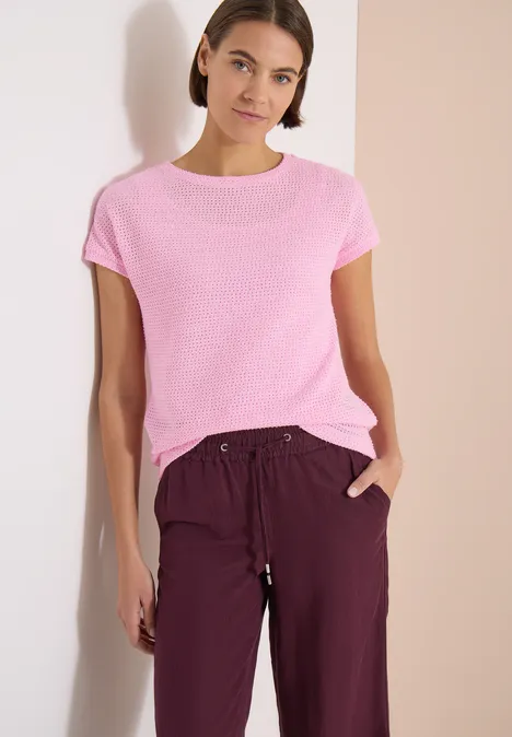 Frau steht vor hellen Wänden, trägt ein pinkes T-Shirt und weinrote, lockere Hose.