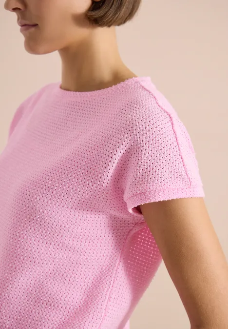 Eine Person in rosa gestricktem Kurzarmshirt vor hellem Hintergrund.