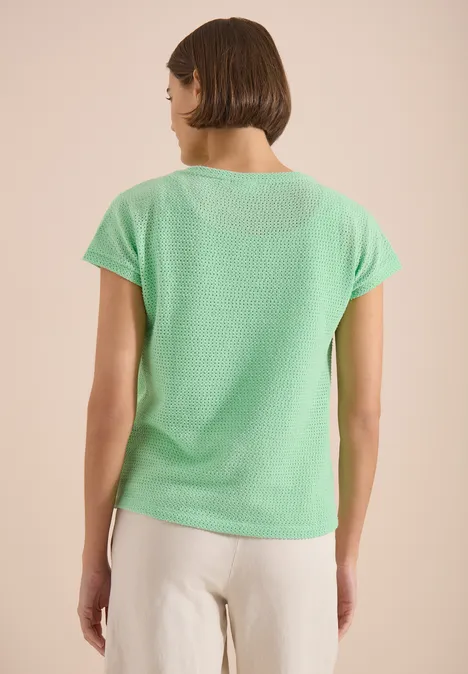 Eine Frau von hinten in grünem T-Shirt und heller Hose vor neutralem Hintergrund.