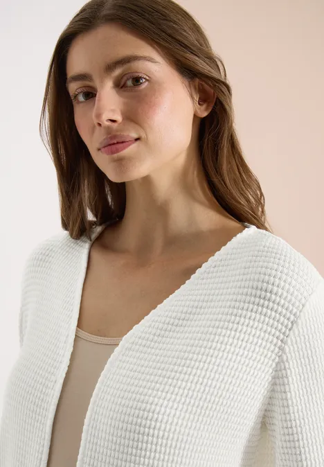 Femme vêtue d'un gilet en maille blanche et d'un débardeur beige, fond beige uni.