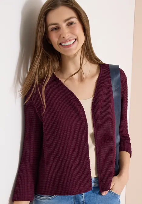 Femme en gilet bordeaux sur un top blanc, main dans la poche, devant un fond clair.