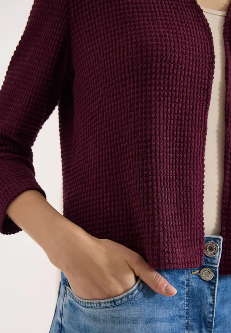 Torse féminin vêtu d’un cardigan bordeaux, d’un haut blanc et d’un jean bleu, main dans la poche