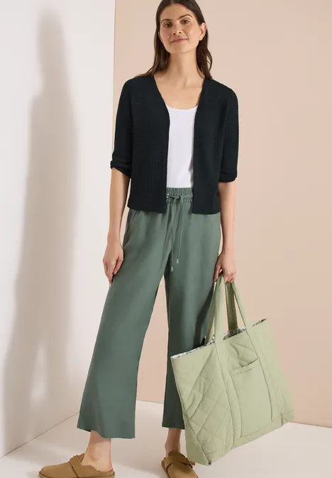 Femme en débardeur blanc, gilet noir et pantalon ample vert, tenant un grand sac matelassé vert clair.