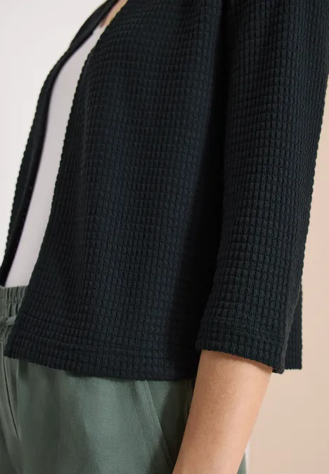 Cardigan noir texturé ample à manches 3/4 porté sur un top blanc et pantalon vert.