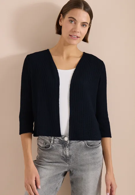 Veste ouverte à manches 3/4 en look ajouré universal blue Femme en cardigan noir court, débardeur blanc et jean gris sur fond beige.
