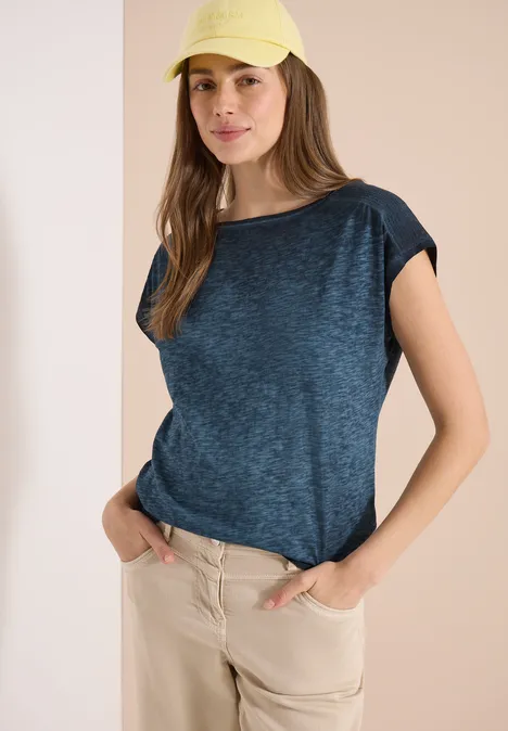 Frau in blauem T-Shirt und beiger Hose vor hellem Hintergrund, Hände in den Taschen.
