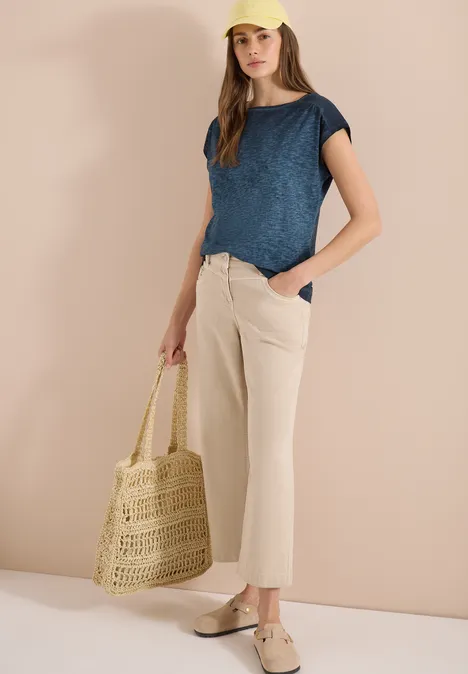 Frau in blauem Shirt, beiger Hose und Espadrilles hält eine Flecht-Tasche vor hellem Hintergrund.