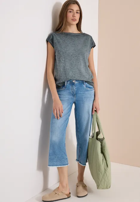 T-Shirt mit Boatneck im Garment-Dye-Look volcanic grey Frau in grauem T-Shirt, hellblauen Cropped-Jeans, beigen Slippern und grünem Steppbeutel vor zweifarbigem Hintergrund.