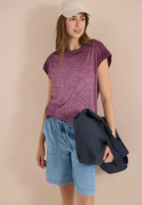 Vrouw in paars T-shirt, jeansshorts en witte pet, met een jas in haar hand.