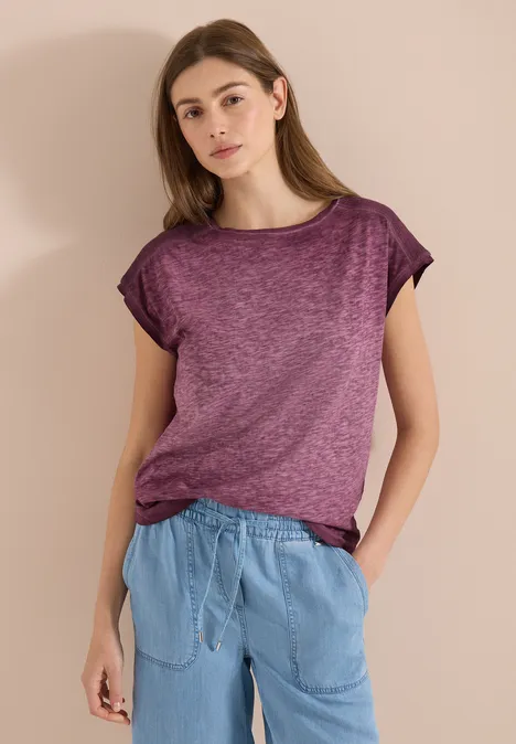 Vrouw draagt paars T-shirt en lichtblauwe broek, ontspannen pose voor een neutrale achtergrond.