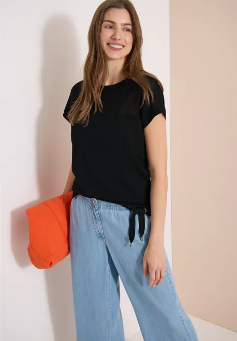 Vrouw draagt zwart T-shirt, lichtblauwe broek en houdt een oranje clutch voor een neutrale achtergrond.