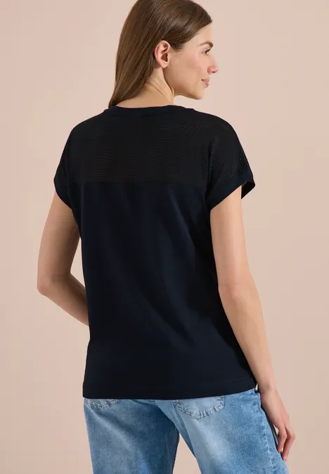 Frau von hinten in dunkelblauem T-Shirt mit kontrastierender Passe und blauen Jeans vor beigem Hintergrund.