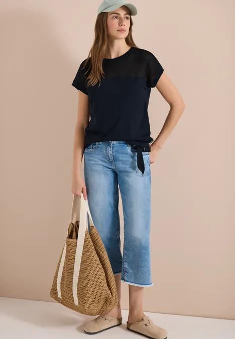 Frau in schwarzem T-Shirt, blauen Jeans, Strohtasche und beigen Hausschuhen vor neutralem Hintergrund.