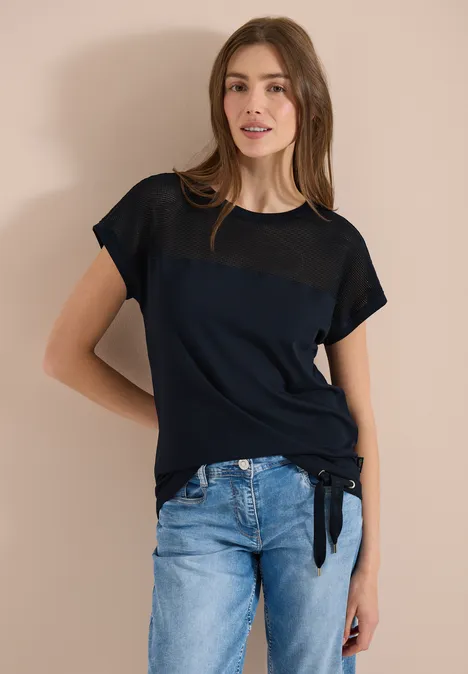 Eine Person in schwarzem T-Shirt mit transparentem Einsatz und blauer Jeans steht vor beiger Wand.