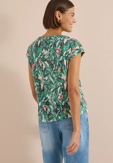 Shirt met burn-out look khaki Vrouw van achteren in een groen shirt met tropisch dessin en spijkerbroek voor een neutrale achtergrond.
