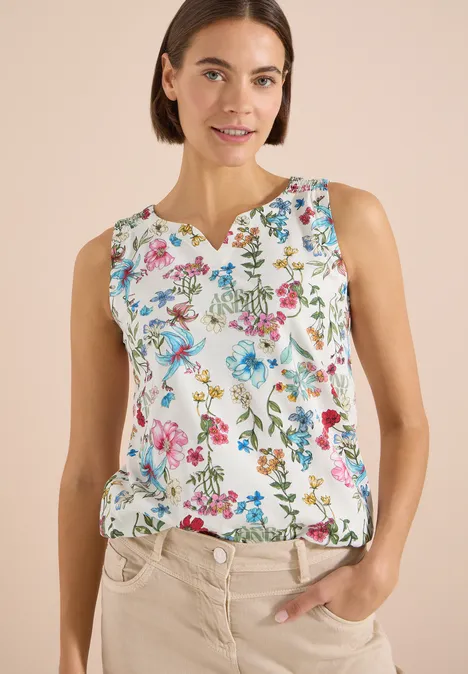Top mit Split Neck und Raffung light alabaster white Eine Frau trägt ein ärmelloses, weißes Top mit buntem Blumenmuster und steht entspannt mit Hand in der Tasche.