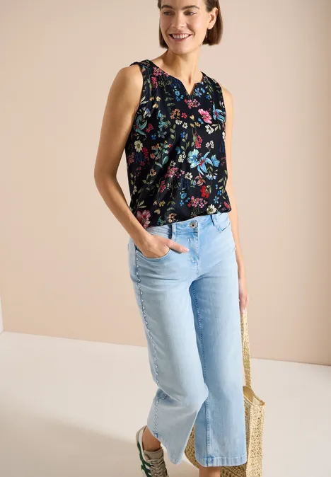 Femme en top sans manches noir à fleurs et jean large bleu clair fendu au bas, tient un sac en paille.