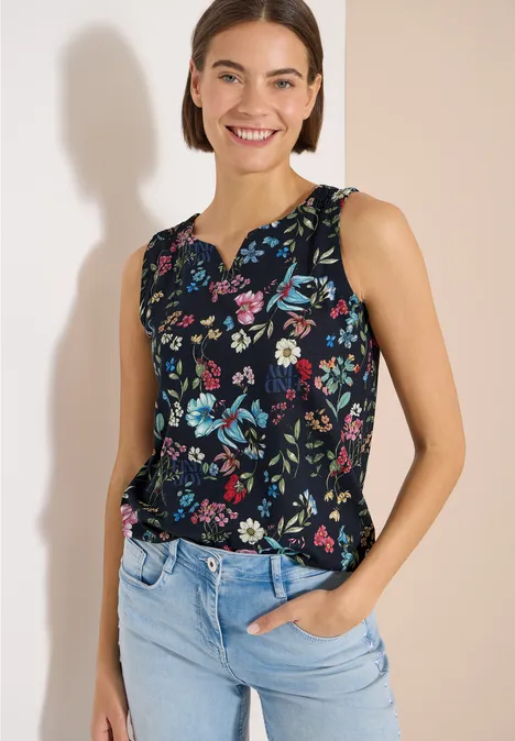 Femme en haut sans manches à motif floral sombre et jean bleu clair devant un mur beige.