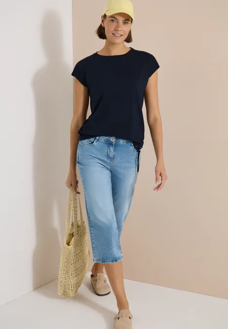 Frau in dunkelblauem T-Shirt, hellblauen Capri-Jeans, hellen Schuhen und gelber Netz-Tasche vor neutralem Hintergrund.