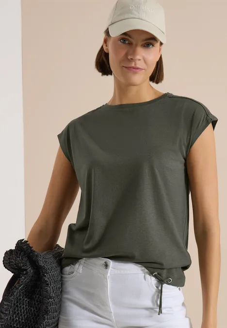 Frau in dunkelgrünem Shirt und hellen Jeans, hält einen schwarzen Pullover vor neutralem Hintergrund.