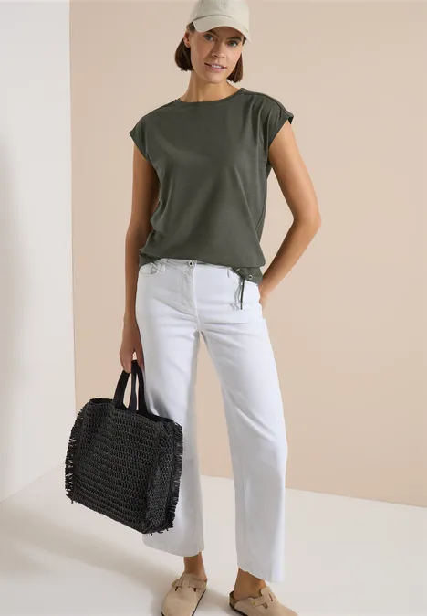 Frau in khakigrünem T-Shirt, weißen Jeans und flachen Sandalen, trägt eine schwarze geflochtene Tasche vor neutralem Hintergrund.