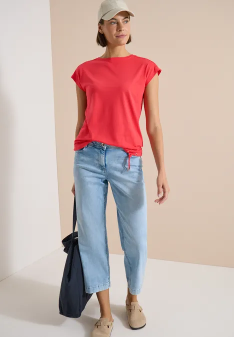 Frau in rotem Shirt, hellblauen Jeans und beigen Schuhen mit dunkler Tasche.