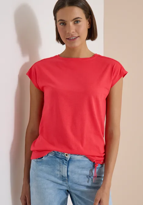 Frau in rotem T-Shirt und blauer Jeans steht vor einer Wand, Gesicht unkenntlich.