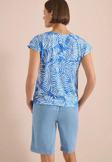 Frau von hinten in blau-weiß gemustertem Shirt und hellblauen Denim-Bermudas vor beigem Hintergrund.