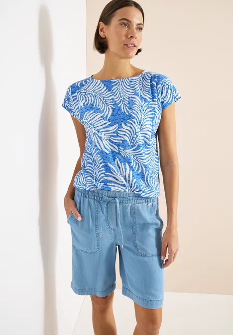 Frau trägt blaues Blattmuster-Shirt und helle Jeans-Bermudashorts vor hellem Hintergrund.