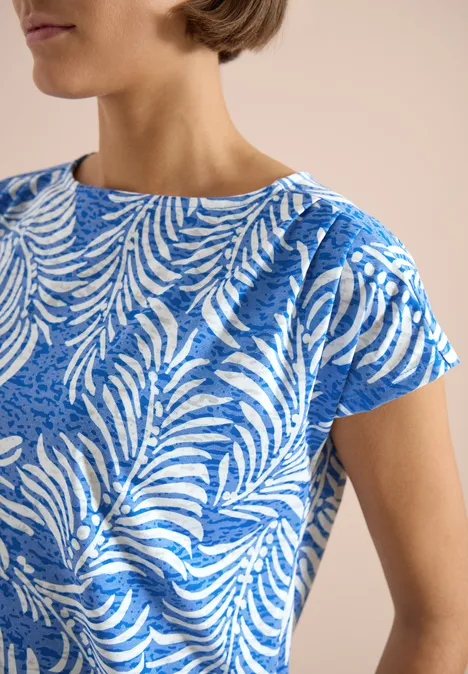 Nahaufnahme eines blauen Kurzarmshirts mit weißem Blattmuster am Oberkörper einer Person.