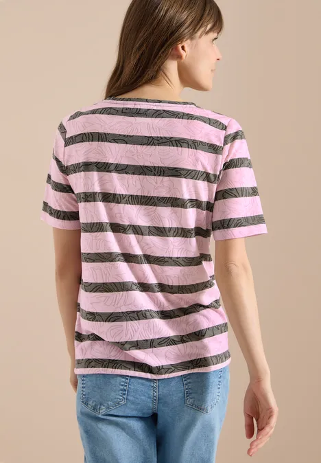 Eine Frau in rosa-braun gestreiftem T-Shirt und Jeans, von hinten vor beiger Wand stehend.