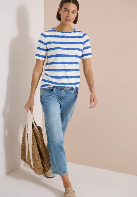 Femme en T-shirt rayé bleu et blanc, jean et sac fourre-tout, marchant devant un mur clair.