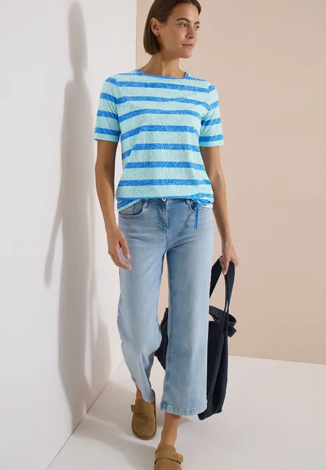 Femme en t-shirt rayé bleu et blanc, jean évasé clair, boots camel et sac bleu marine devant un mur clair.