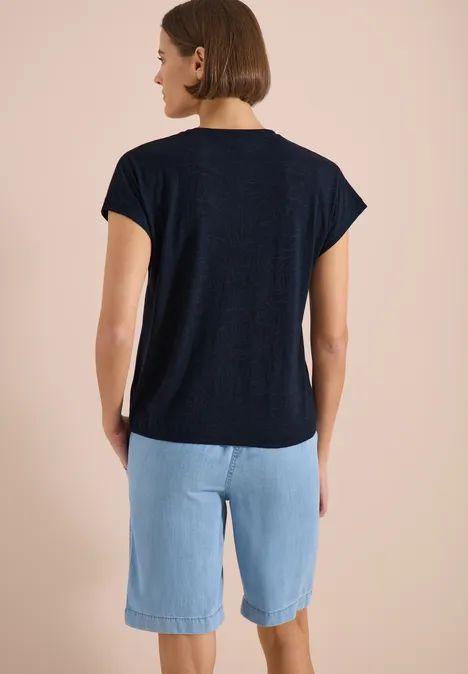 Eine Frau von hinten, trägt ein dunkelblaues T-Shirt und hellblaue knielange Shorts vor neutralem Hintergrund.