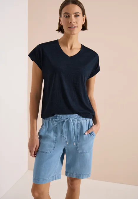 Frau in schwarzem V-Ausschnitt-Shirt und hellblauen Jeans-Shorts mit Taschen vor neutralem Hintergrund.