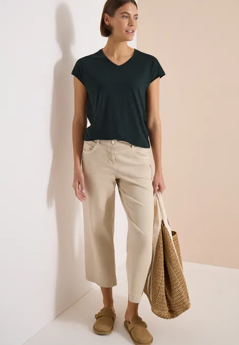 Frau in schwarzem T-Shirt mit V-Ausschnitt, beigen weiten 3/4-Hosen, Basttasche und flachen Mokassins vor hellem Hintergrund.