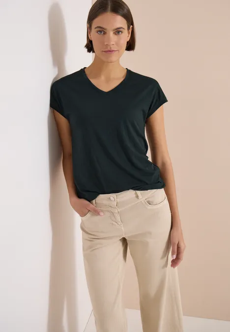 Frau lehnt entspannt an heller Wand, trägt schwarzes T-Shirt und beige Hose.