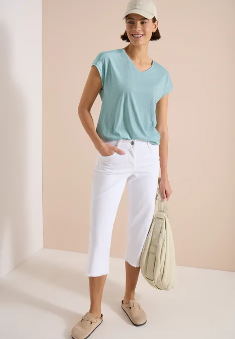 Frau trägt mintgrünes T-Shirt, weiße Caprihose, beige Slipper und hält eine helle Tasche.