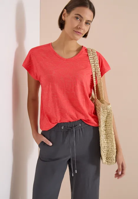 Shirt met ton-sur-ton burn-outpatroon coral Frau mit rotem T-Shirt, dunklen Stoffhosen und einer Strohtasche vor hellem Hintergrund.