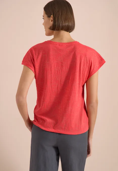Shirt met ton-sur-ton burn-outpatroon coral Rückansicht einer Person in rotem T-Shirt und grauer Hose, seitlich leicht zum Betrachter gedreht.