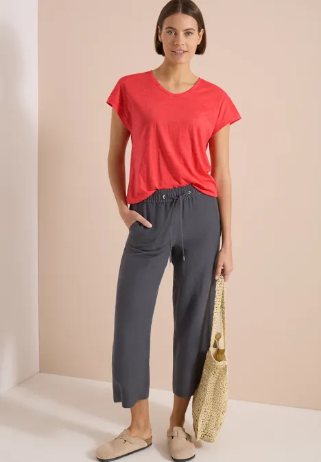 Shirt met ton-sur-ton burn-outpatroon coral Frau in rotem T-Shirt und grauen, knöchellangen Jogginghosen mit Schnürbund, trägt Basttasche und Espadrilles.